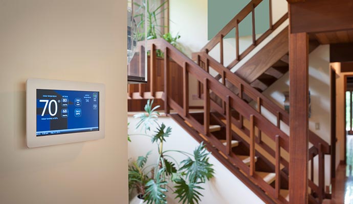 Smart thermostat mounted on a wall near a wooden staircase with indoor plants, displaying 70 degrees Fahrenheit